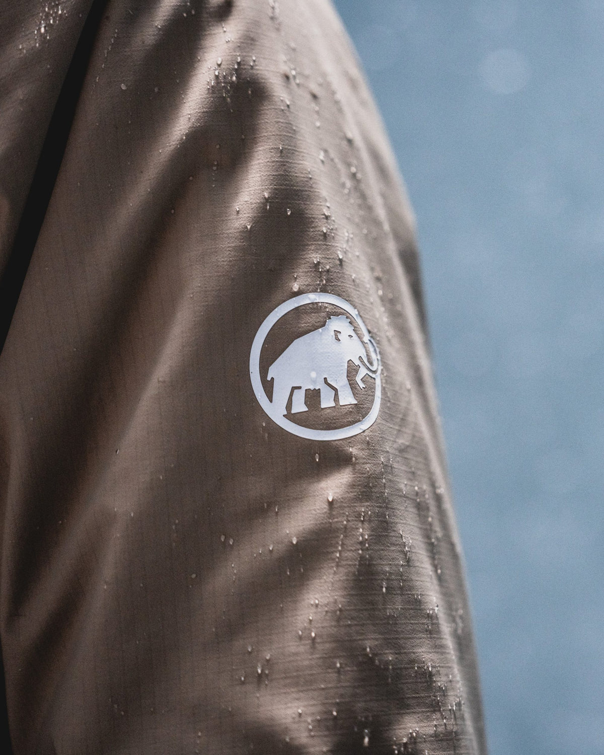 Close-up of a Mammut brown jacket sleeve with water droplets and the iconic white mammoth logo inside a circle.