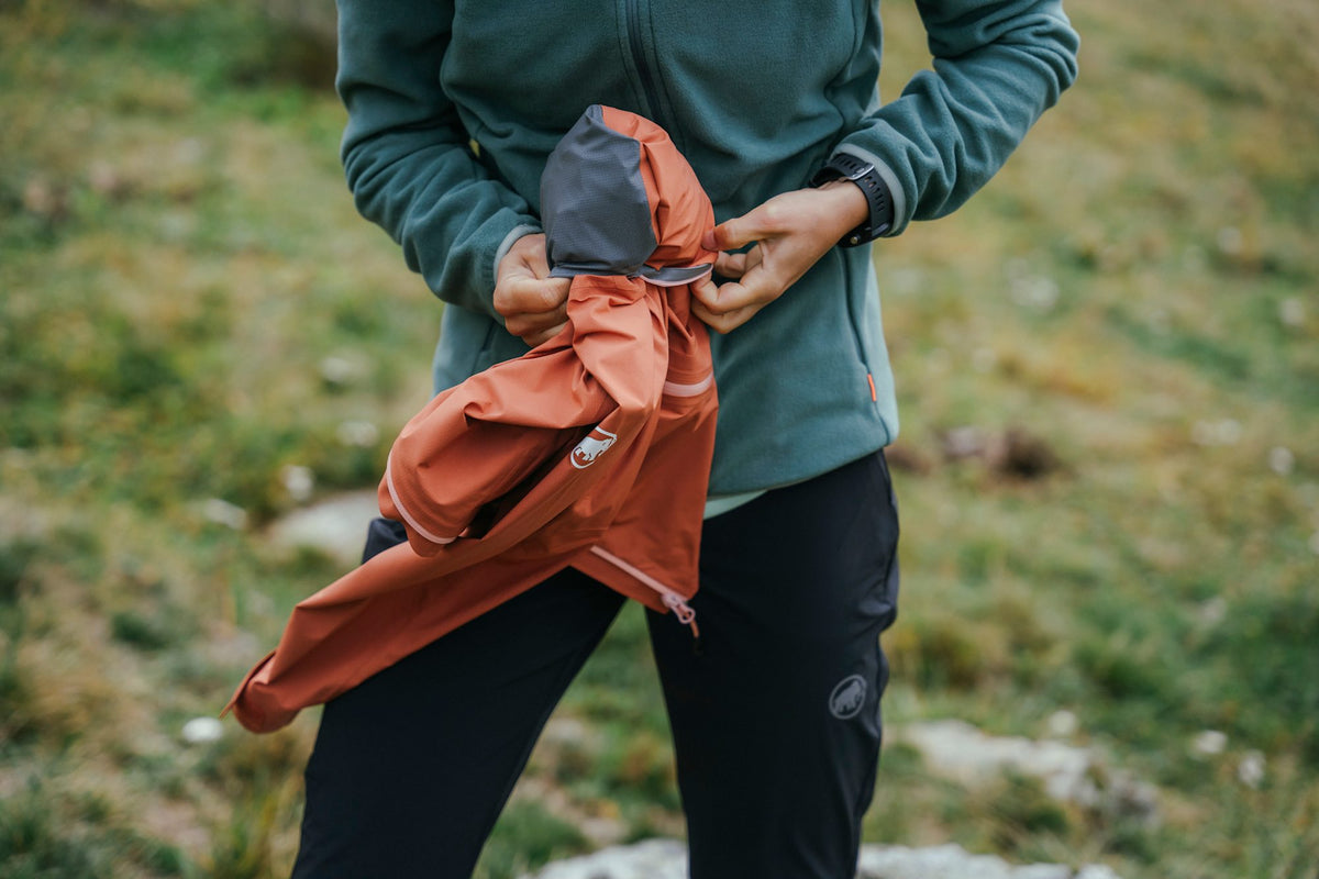 Person in Mammut outdoor gear compressing an orange jacket, standing on a grassy terrain.