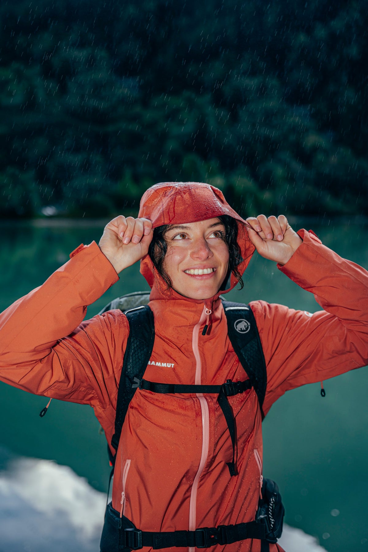 Person smiling in a Mammut orange rain jacket, adjusting the hood outdoors near a scenic lake and lush greenery in the background.