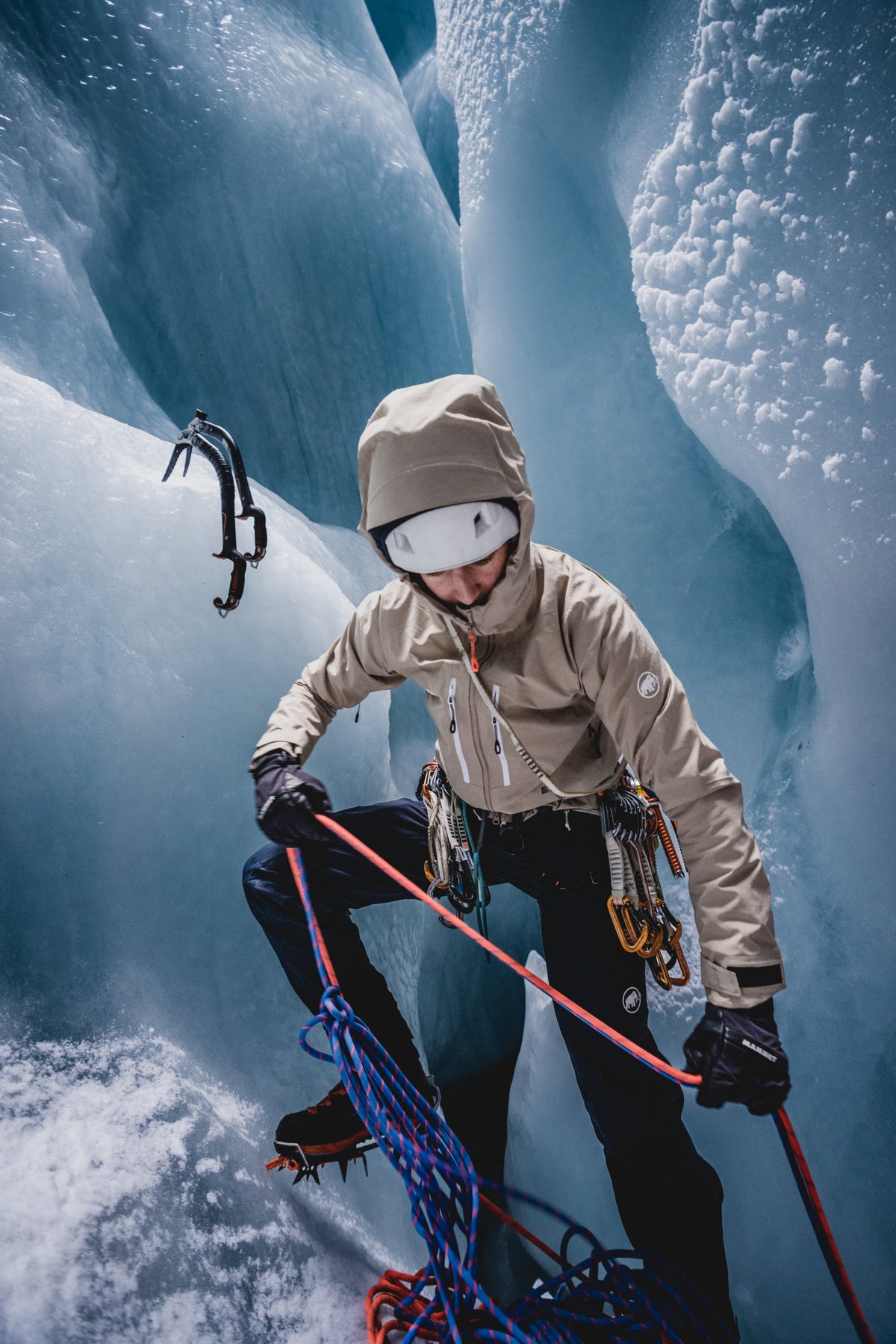 Mammut climber in specialized winter gear ascending icy terrain with ropes and ice axes, amidst striking blue ice formations.