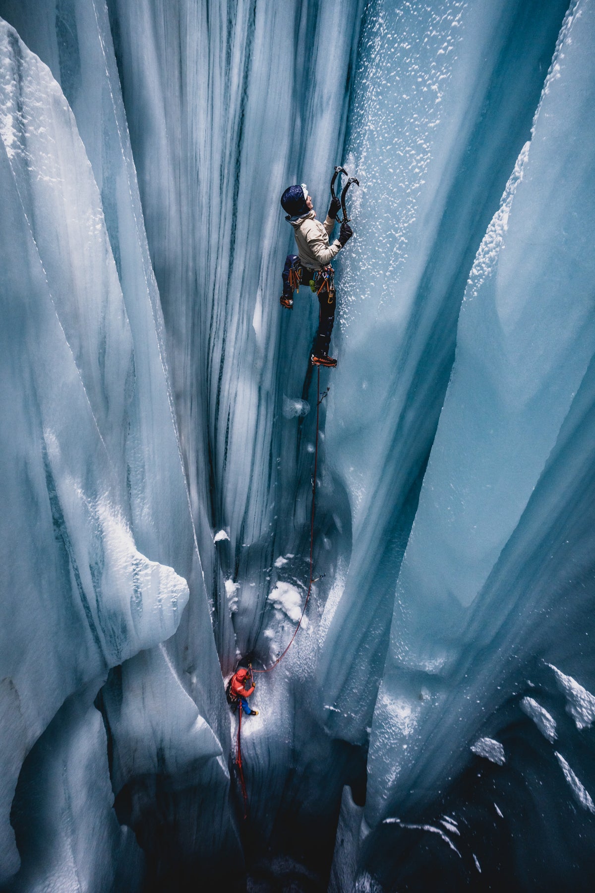 Two climbers equipped with Mammut gear ascend a towering icy crevasse; one higher up uses ice axes, roped for safety, surrounded by stunning blue ice walls.
