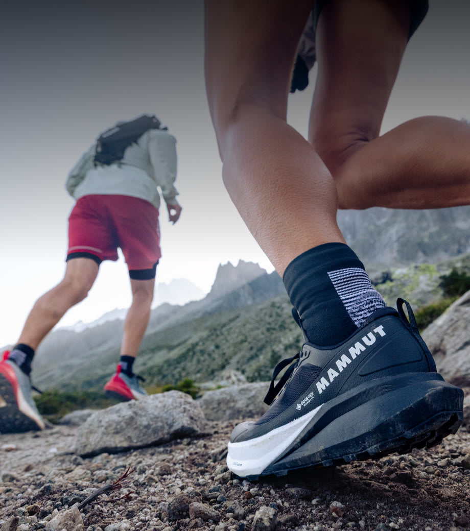 Two athletes trail running on rocky mountain terrain, highlighting black Mammut trail running shoes and alpine landscape.
